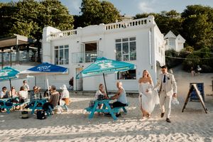 Bride and groom walk hand-in-hand on sandy beach near white seaside venue with blue umbrellas and guests.