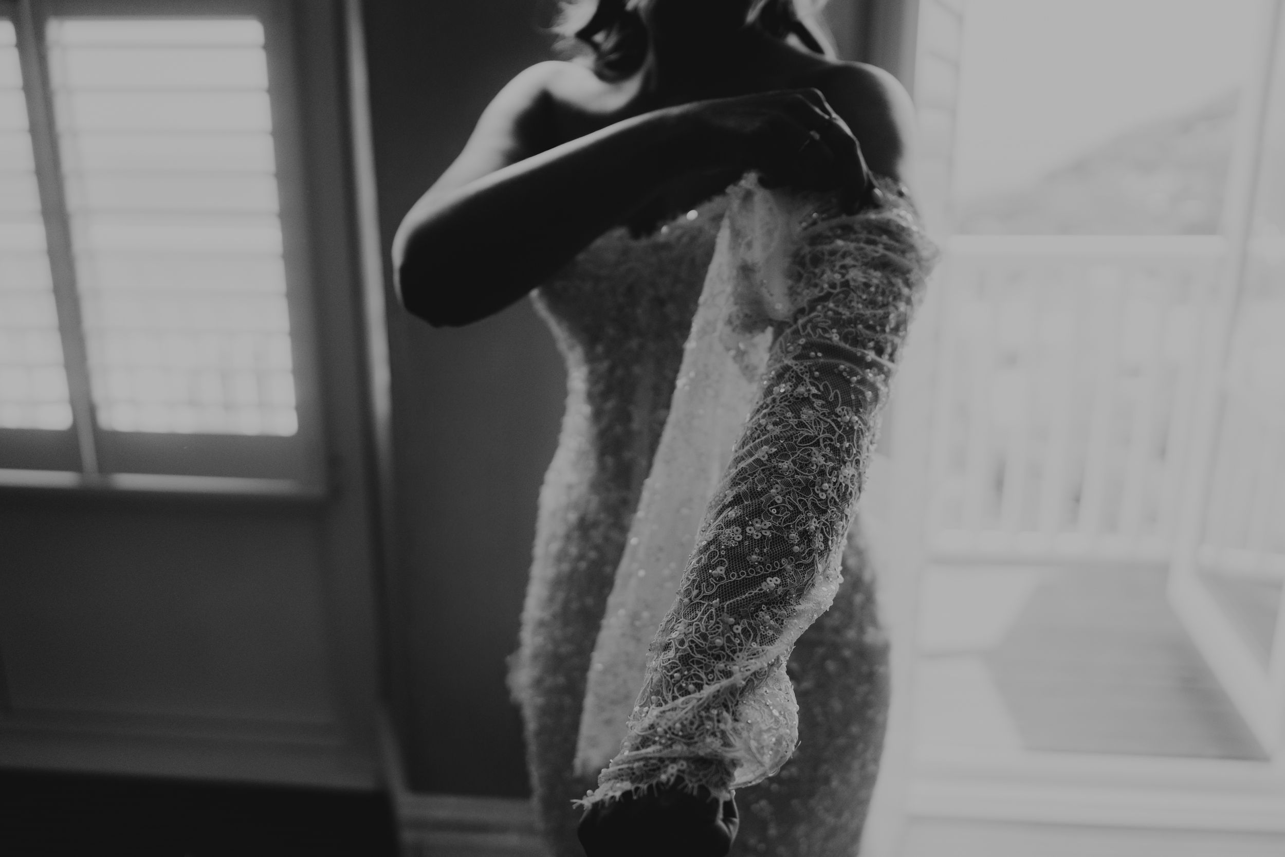 Black and white photo of a bride in a beaded gown holding her veil near a window.