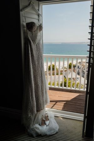 Wedding dress hanging in balcony doorway with ocean view, paired with bridal heels, in a coastal hotel room.