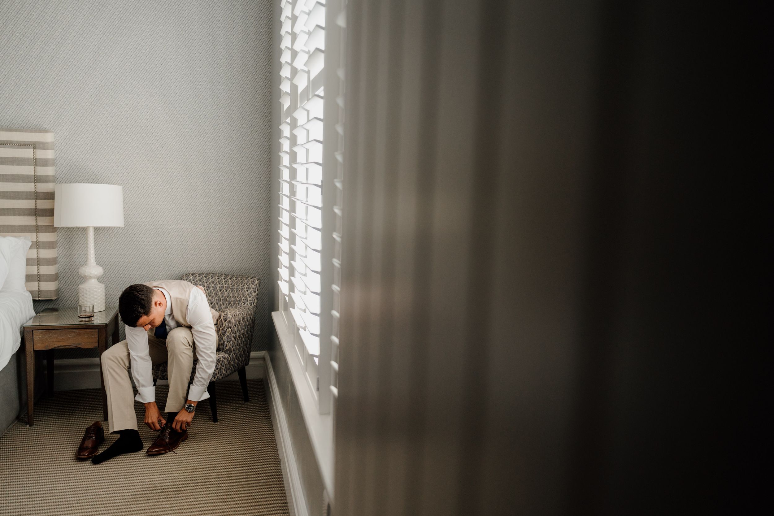 Groom in a white suit tying his dress shoes beside a sunlit window with plantation shutters on his wedding day.