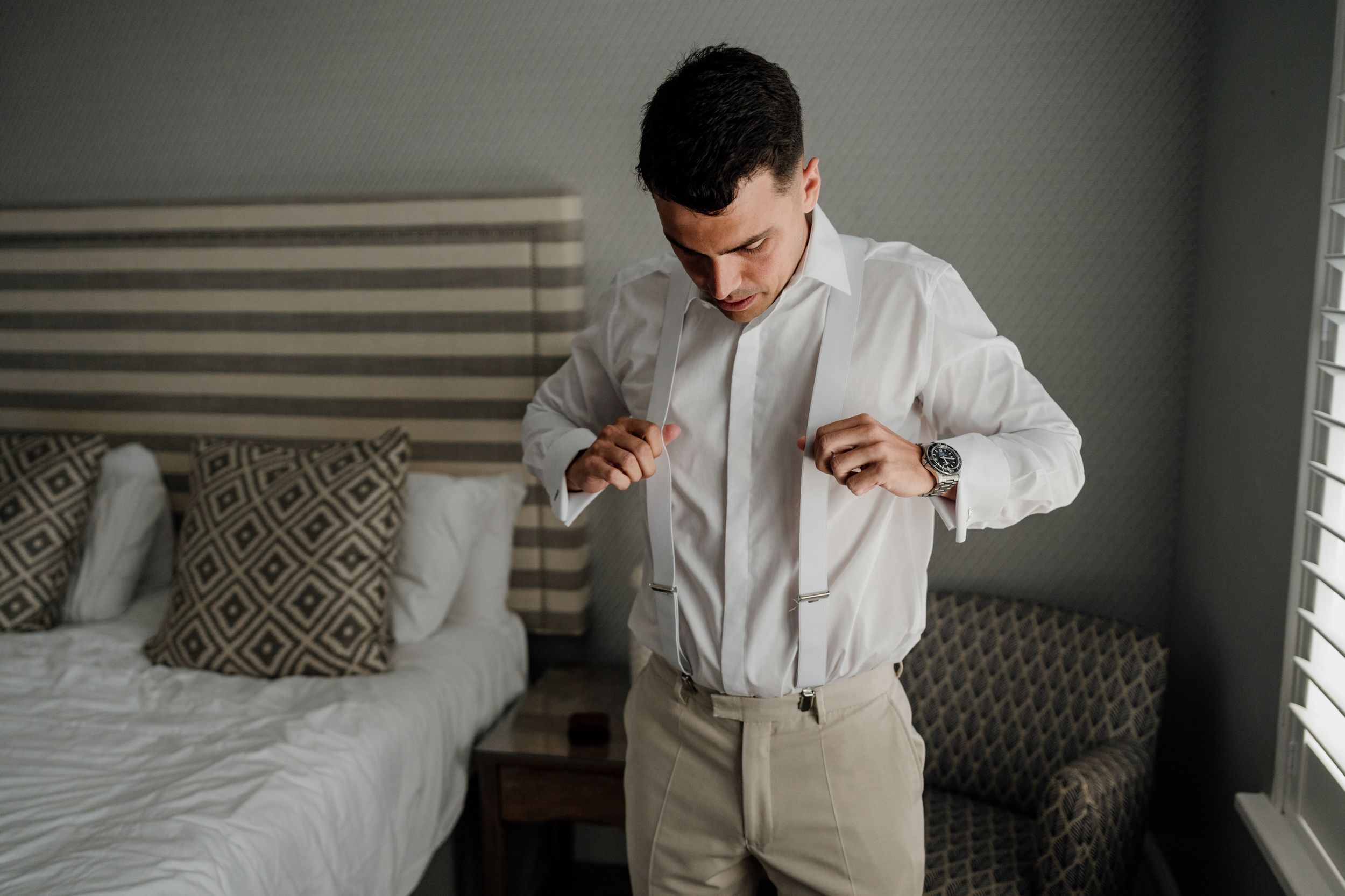 A groom in a white dress shirt and khaki pants buttoning his shirt while getting ready in a hotel bedroom.