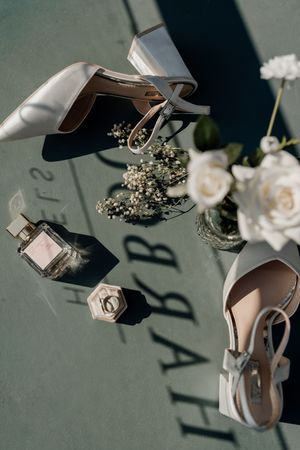 Elegant bridal flat lay featuring white heels, pearl ring, perfume bottle, baby's breath, and white roses on a green surface.