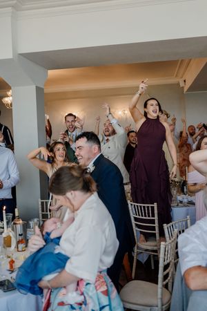 Wedding guests celebrate joyfully at a reception, with a woman in burgundy dress standing on chair, arms raised in excitement.