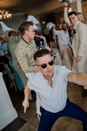 Man in sunglasses takes selfie while dancing at lively wedding reception with guests celebrating in background.