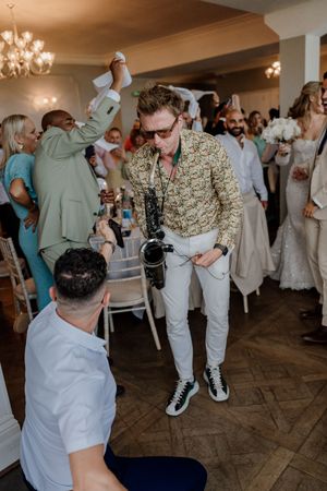 Photographer with camera around neck dancing at a wedding reception in an elegant venue with guests celebrating.