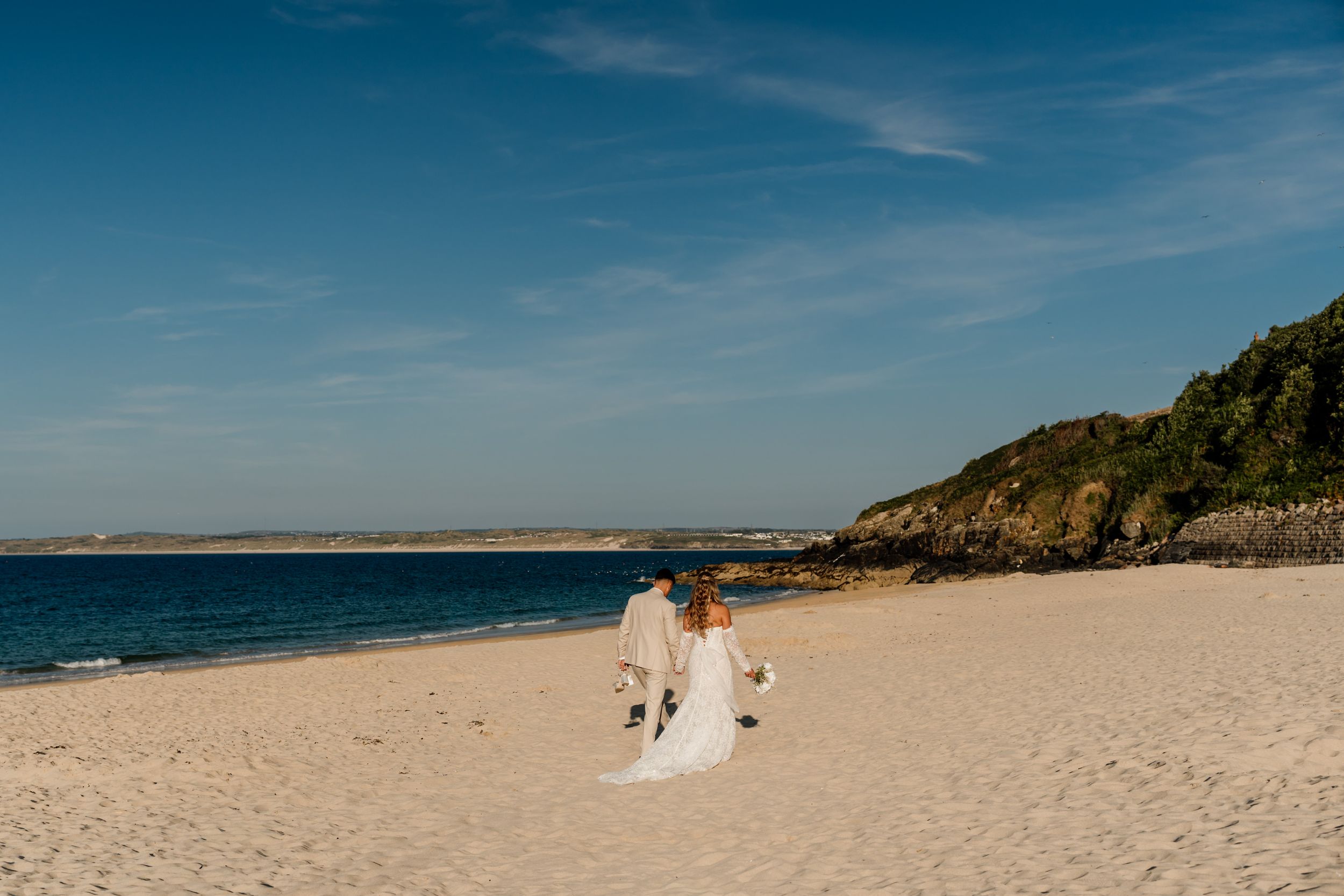 Bride and groom walking hand in hand along a sandy beach with turquoise water and rocky headland under blue skies.