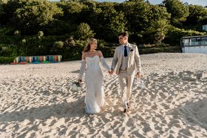Bride and groom walk hand in hand on a sandy beach, bride in white gown, groom in tan suit.