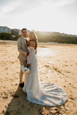 Bride in lace gown and groom in tan suit pose on sandy beach at golden sunset with coastal town backdrop.
