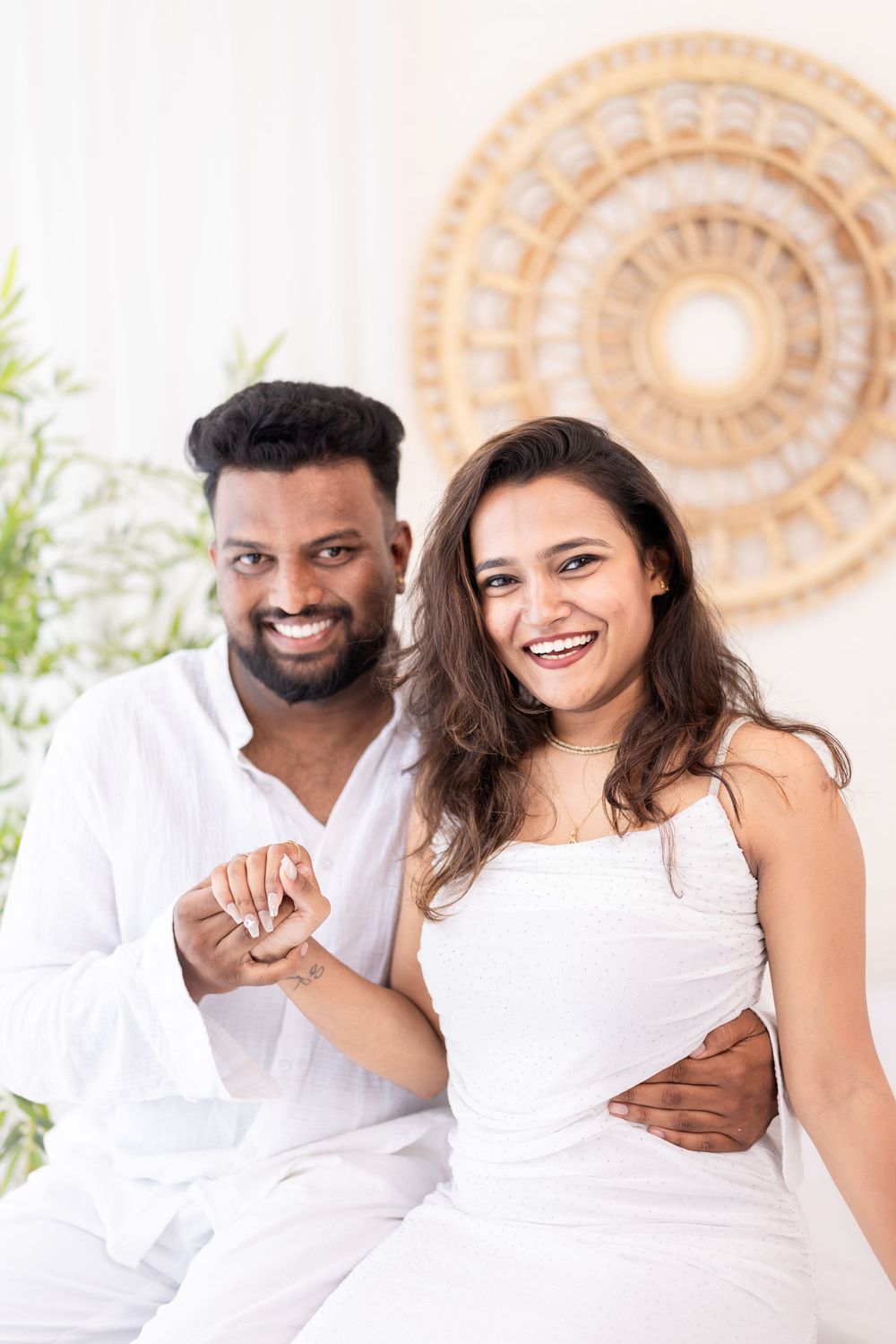 Happy Indian couple in white outfits smiling and holding hands, sitting together in a bright modern room.