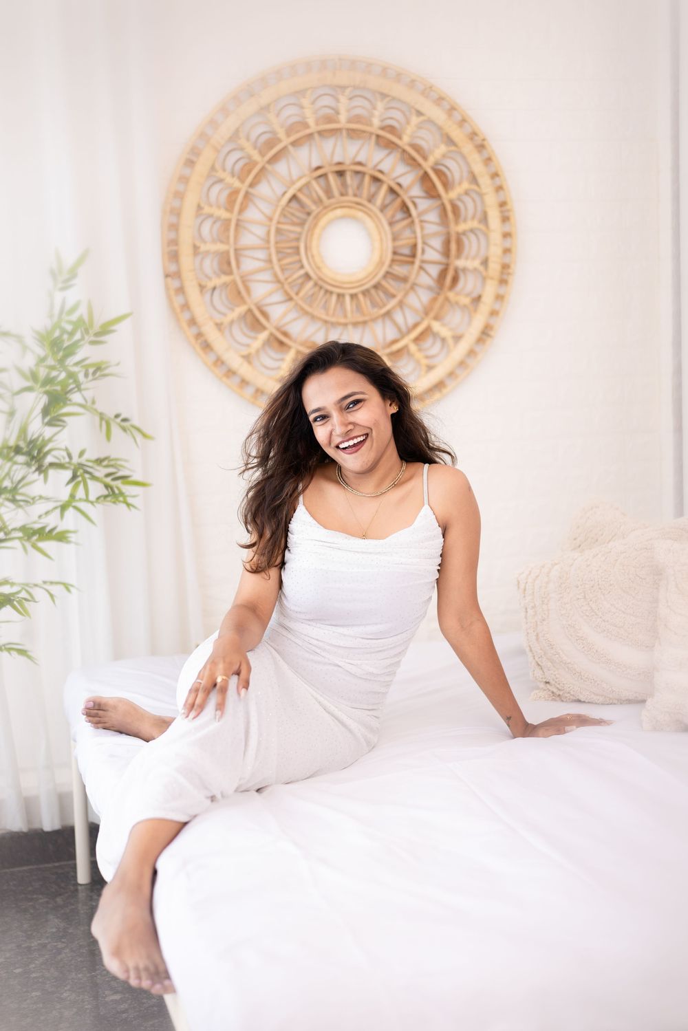 Smiling woman in white outfit sitting on a bed with a decorative mandala wall art and plant in a bright room.