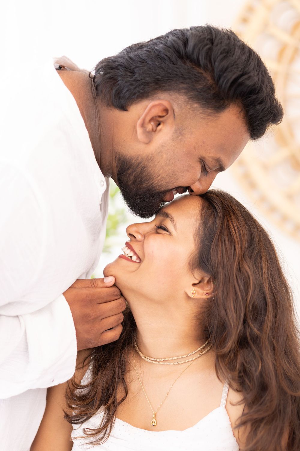 A man tenderly kisses a smiling woman on the forehead while holding her chin in a romantic indoor setting.