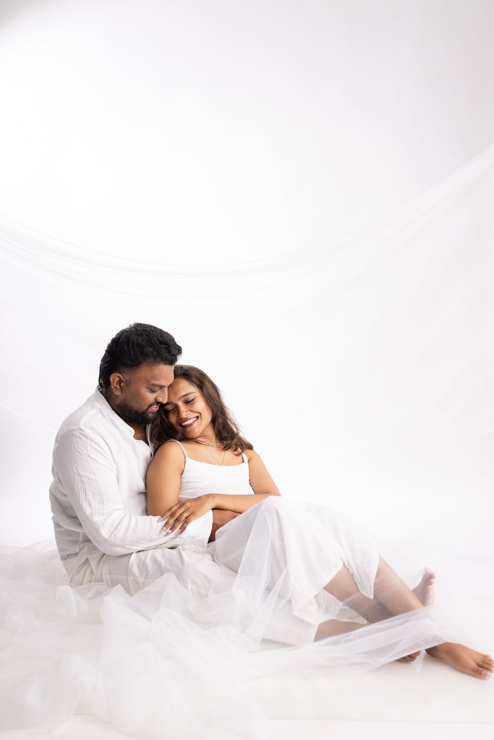 A couple in white outfits sit together, the man embracing a smiling pregnant woman against a white background.