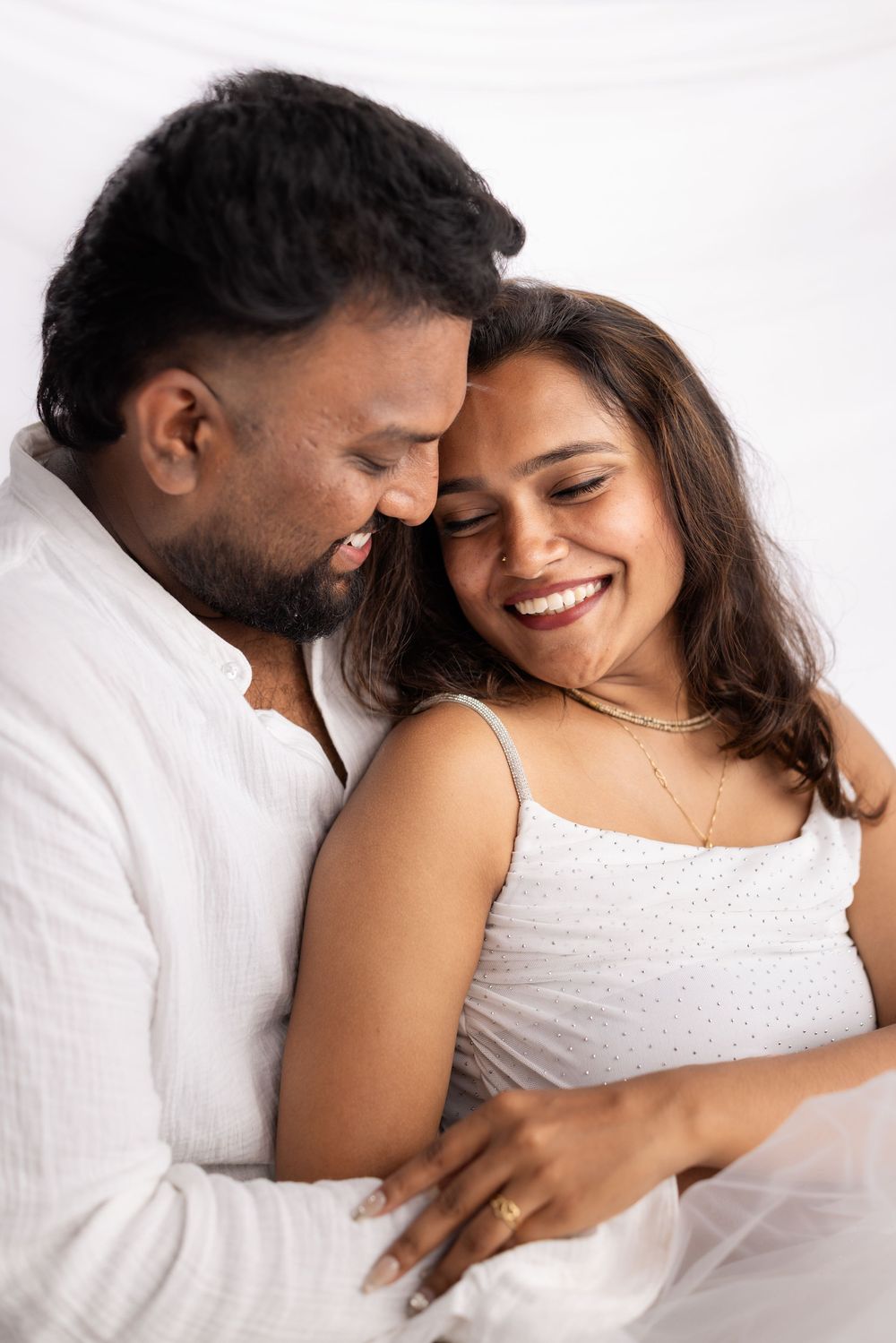 A happy couple in white clothing share a tender embrace, smiling together against a soft white background.