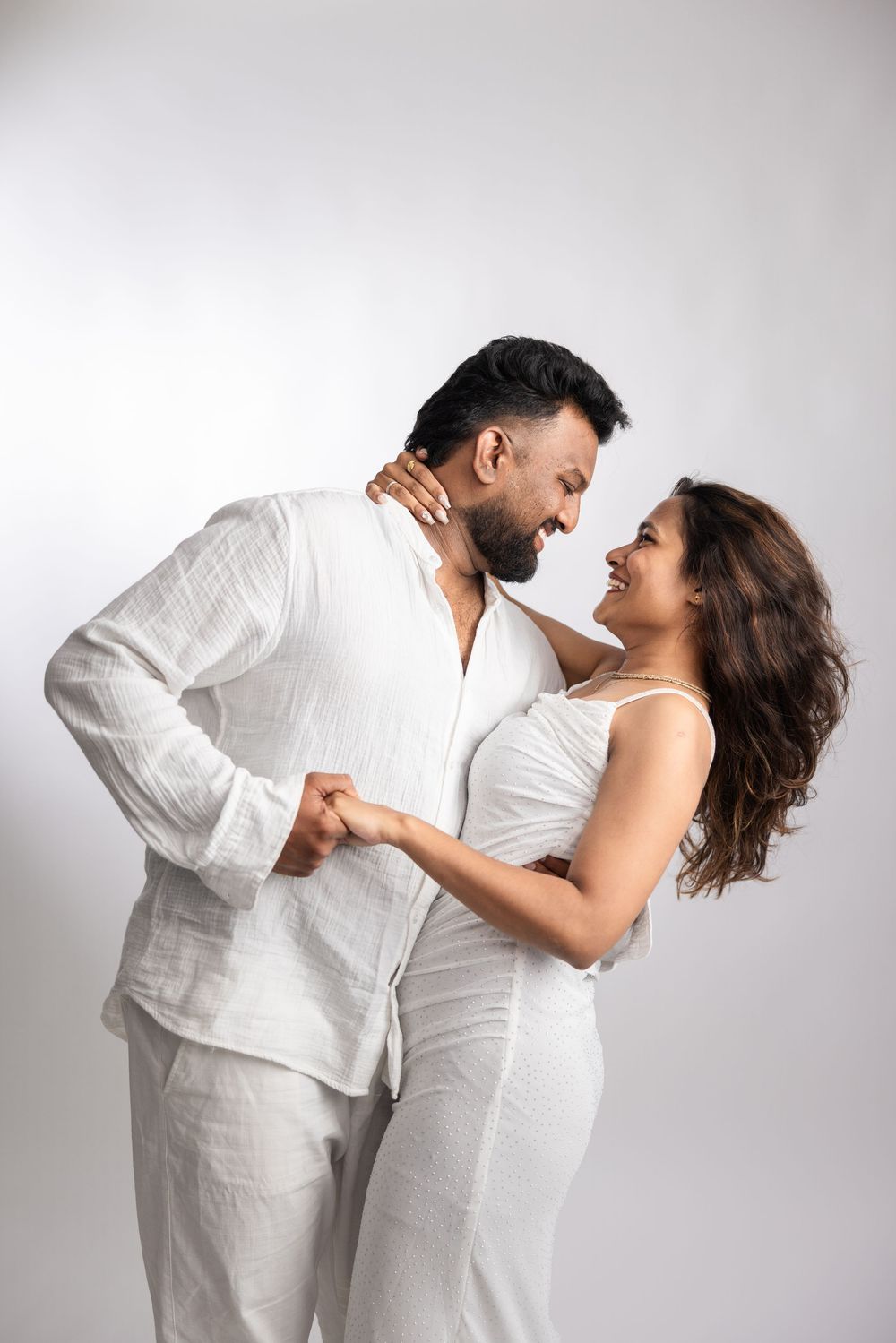 Happy couple in matching white outfits sharing a romantic moment, smiling at each other against a white background.