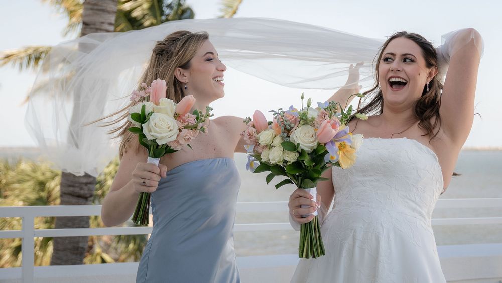 Bride and bridesmaid laughing outdoors holding floral bouquets with veil flowing in the wind at a seaside wedding.