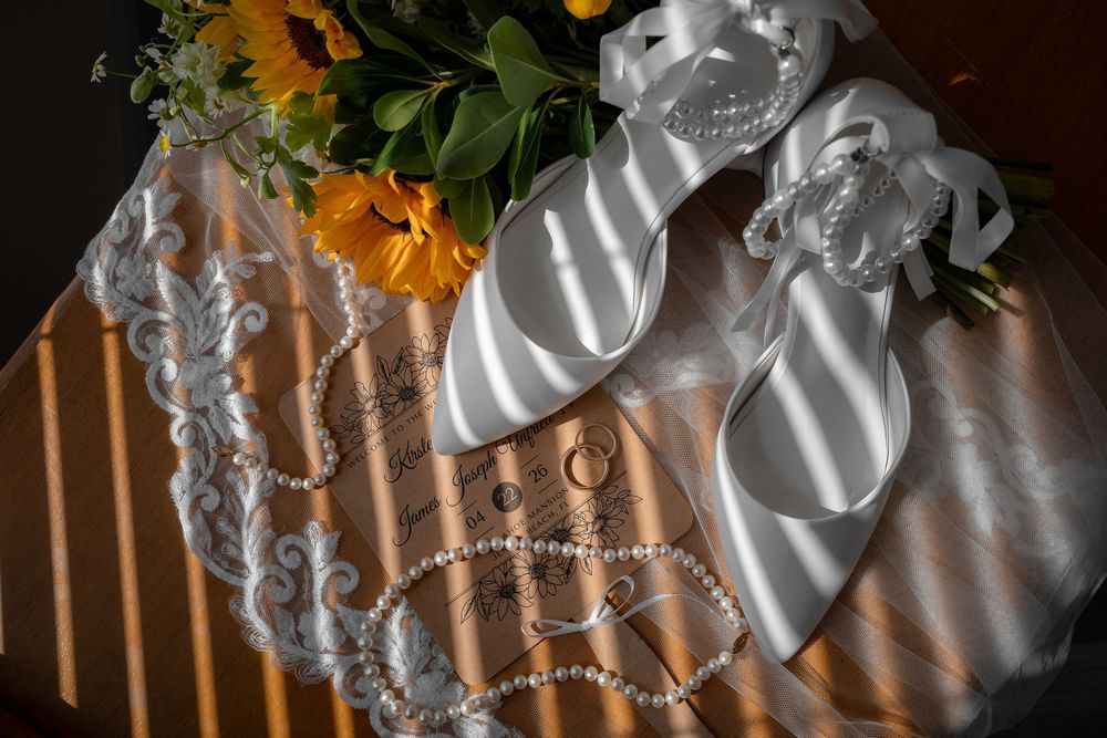 White satin bridal heels with ribbon ties rest on lace fabric beside pearl necklace and sunflower bouquet.