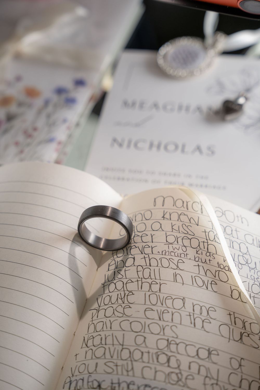 A silver wedding band rests on an open journal with handwritten vows, beside a Meaghan and Nicholas wedding invitation.
