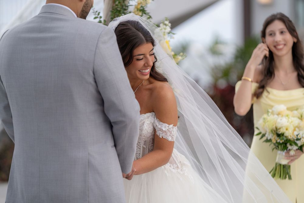 Bride in lace off-shoulder gown laughs joyfully at altar while bridesmaid in yellow dress holds bouquet nearby.