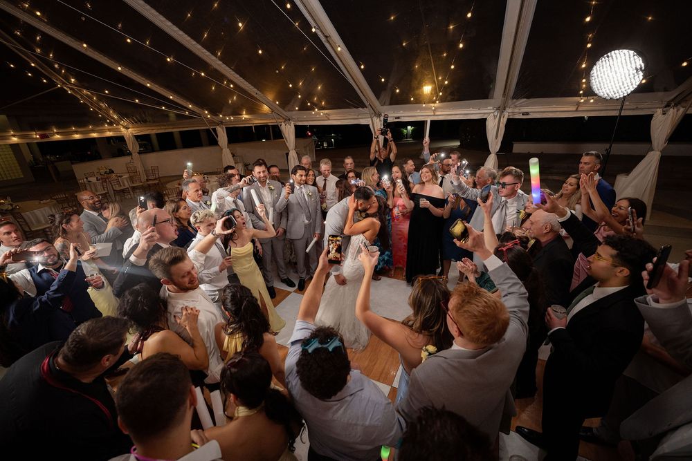 Joyful wedding reception crowd dancing under string lights in a tent venue.