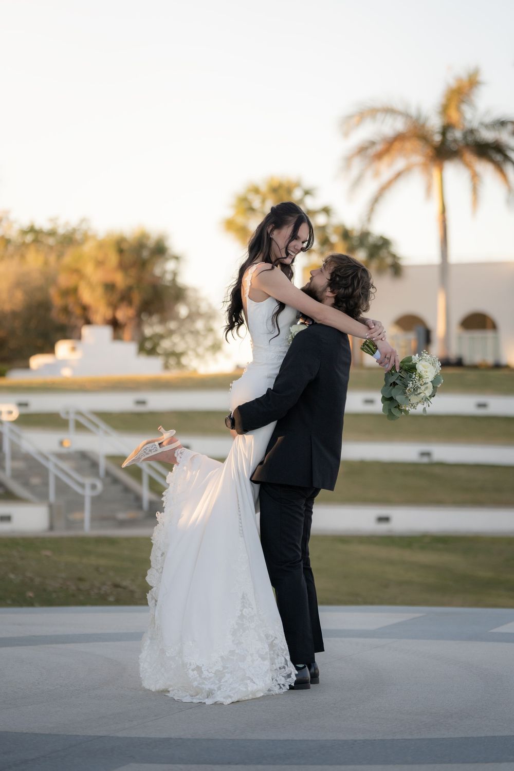 Groom lifts bride in wedding attire outdoors, palm trees and white building in golden hour background.