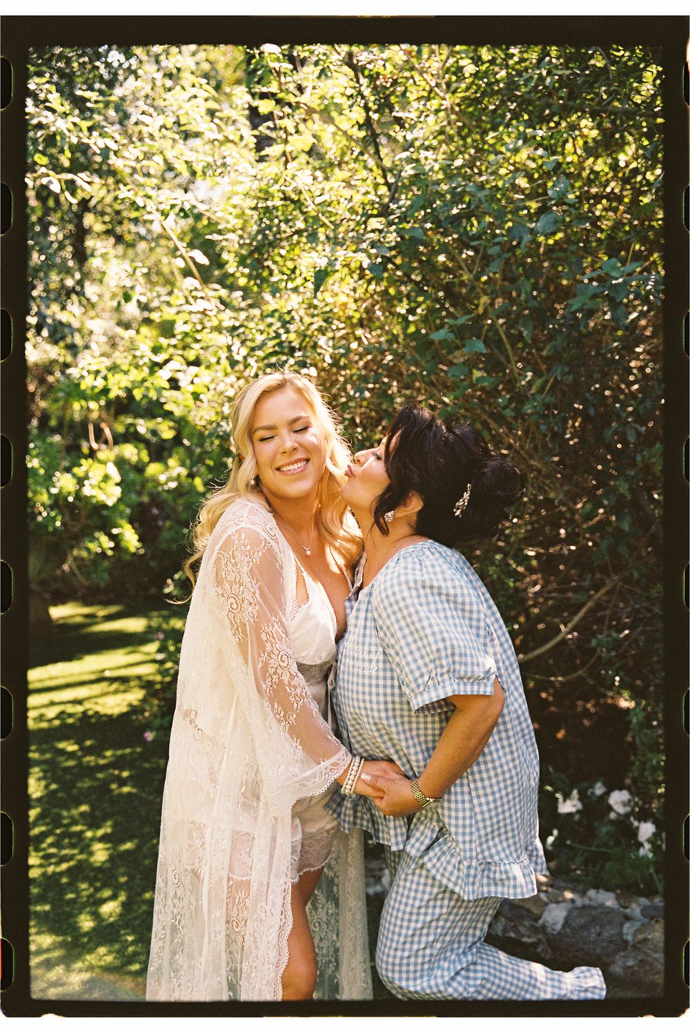 Two women embrace and laugh together in a sunlit garden, one wearing a lace robe and the other a plaid dress.