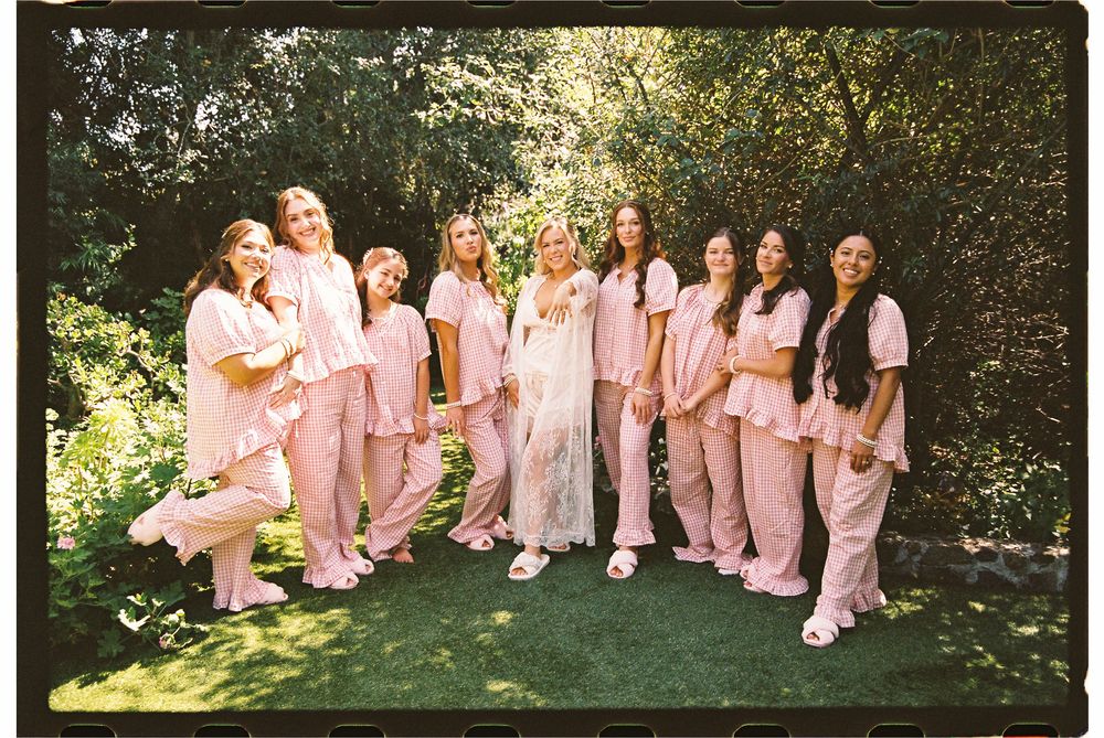 Group of women in matching pink pajamas posing outdoors in a lush garden setting for a bridal party photo.