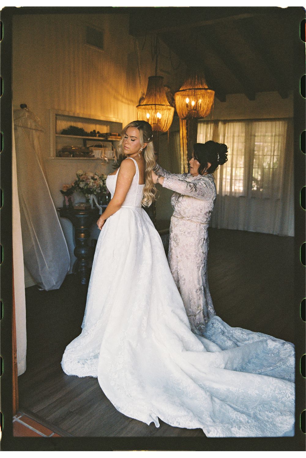A bride in a white lace gown gets her dress fastened by a woman in a lace dress inside a warmly lit room.