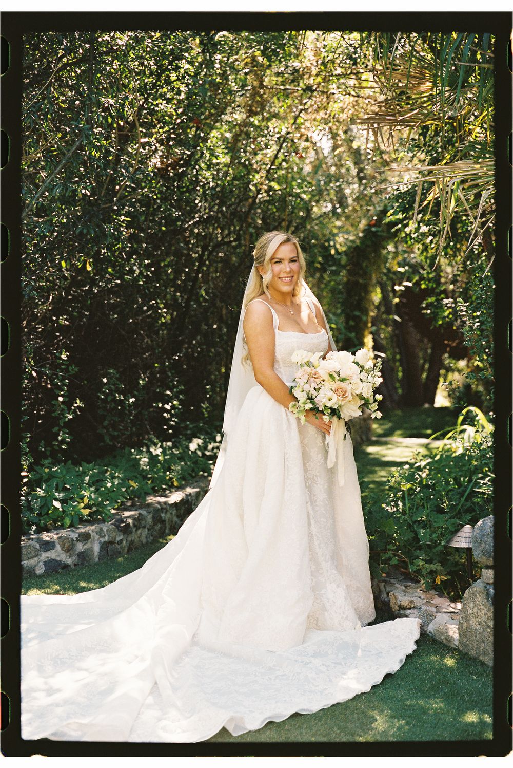 Radiant bride in a flowing white gown with long train holds a bouquet in a lush garden setting.