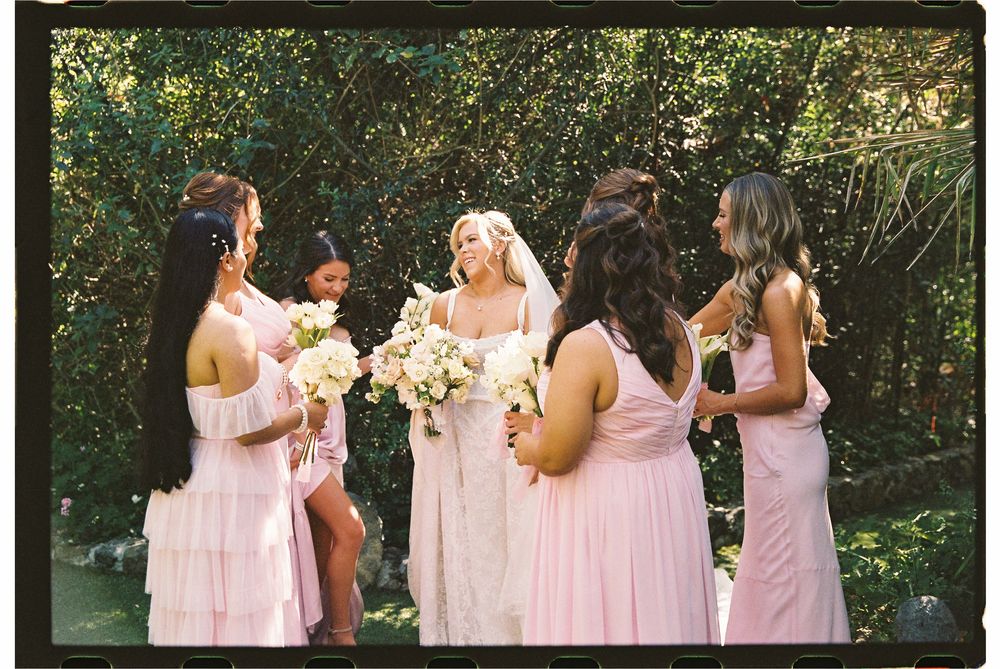 Bride in white gown and veil with bridesmaids in pink dresses holding bouquets outdoors.
