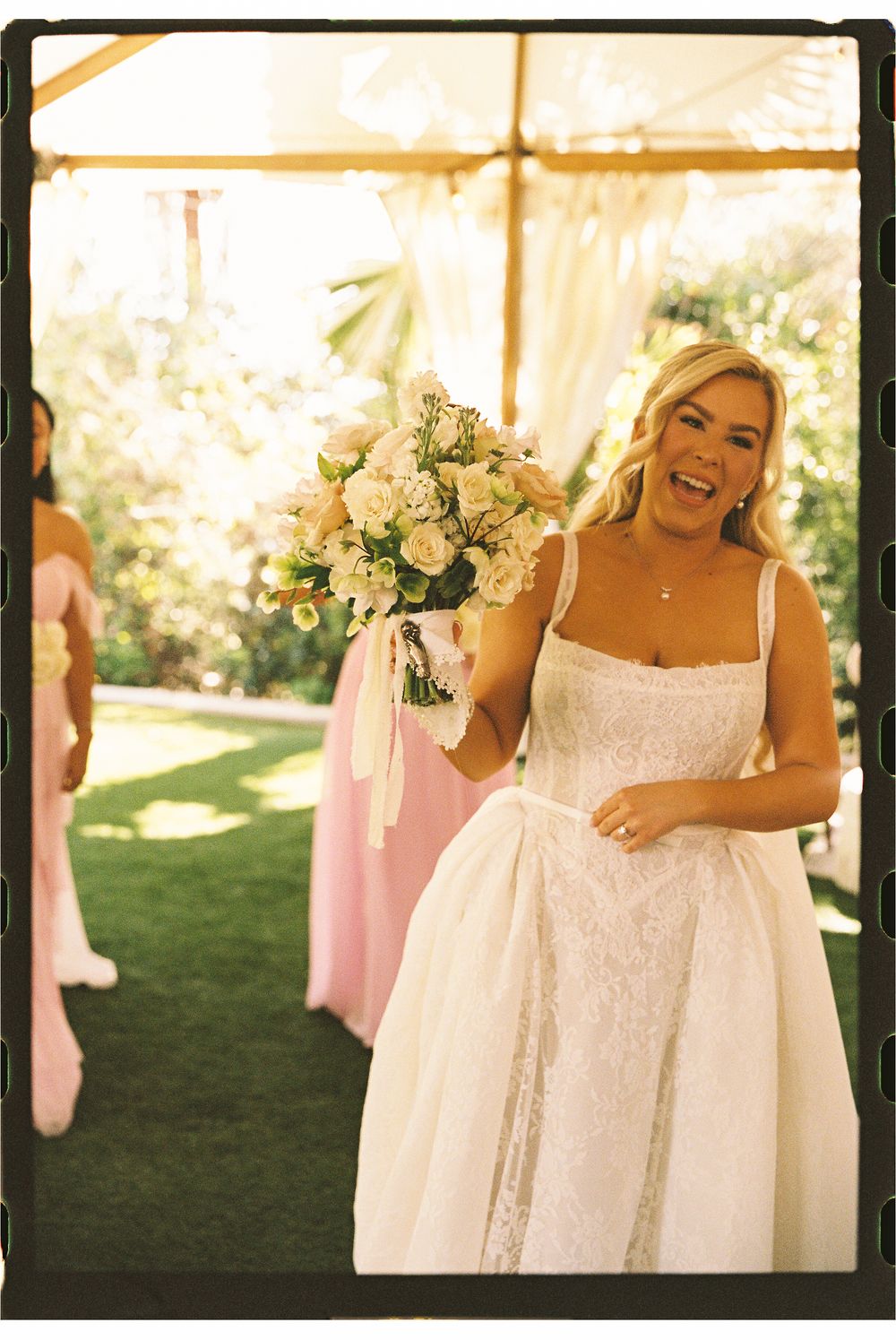 Smiling bride in white lace wedding dress holding bouquet of white roses with bridesmaids in pink dresses.