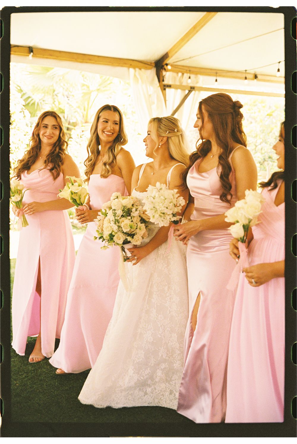 Bride with bridesmaids in pink satin gowns holding white bouquets under a tent at a wedding.