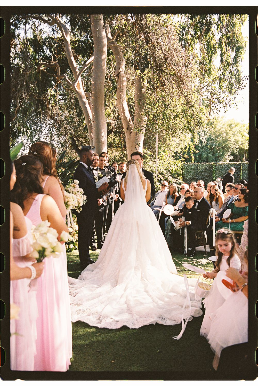 Outdoor wedding ceremony with bride in flowing white gown, pink bridesmaids, and guests seated among tall trees.