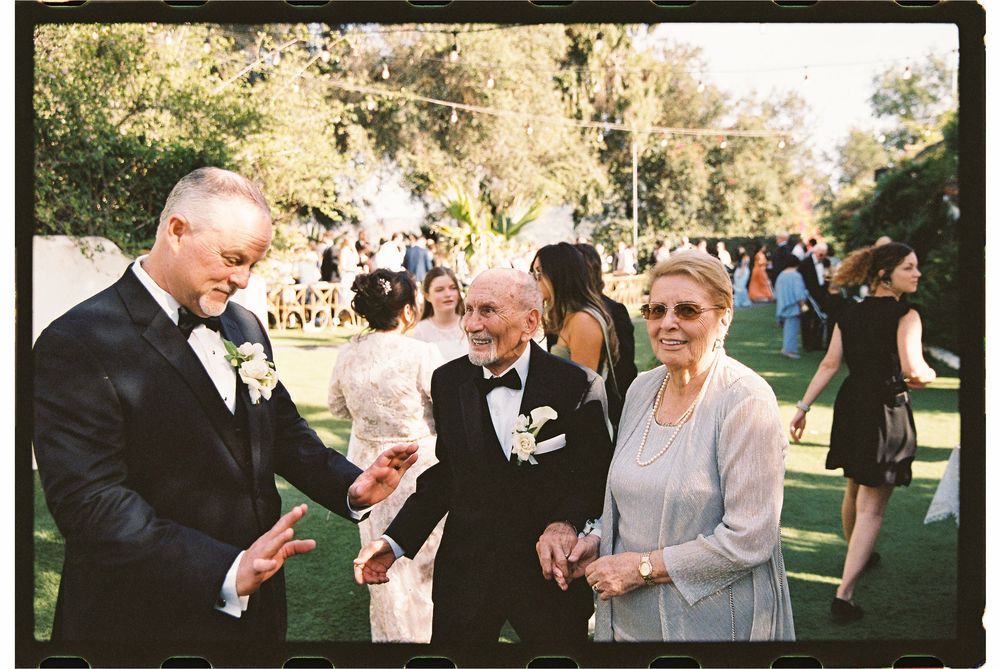 Wedding guests in formal attire socialize and laugh outdoors on a sunny lawn during a wedding reception.