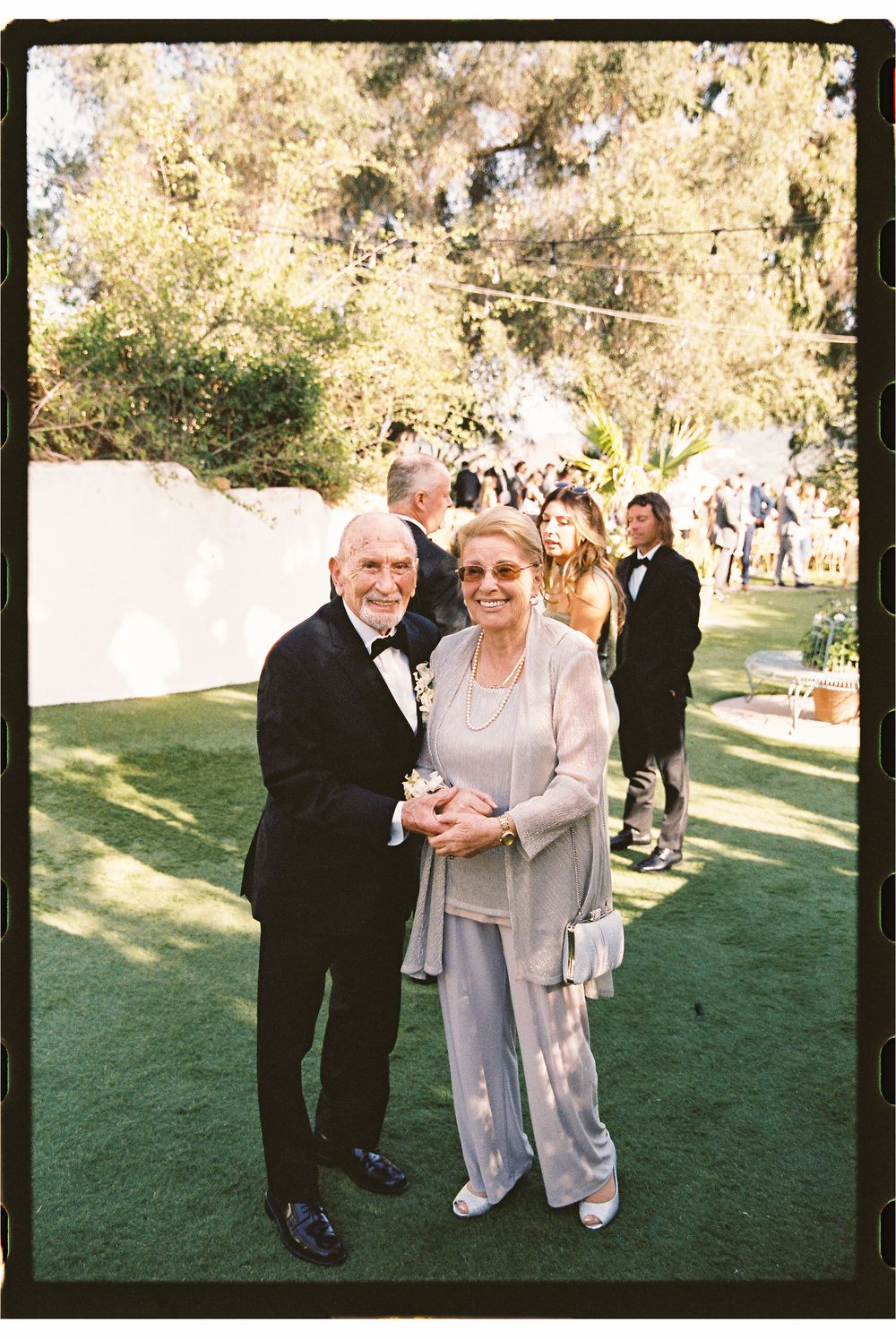 Elderly couple dressed formally smile together on a green lawn at an outdoor wedding reception.
