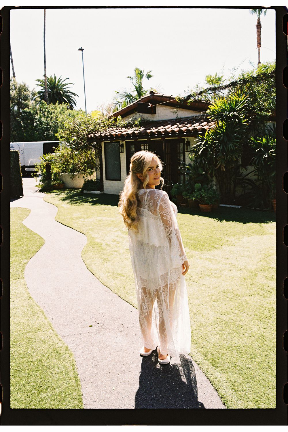 Woman in sheer white lace maxi dress standing on garden path outside Mediterranean-style home with lush greenery.