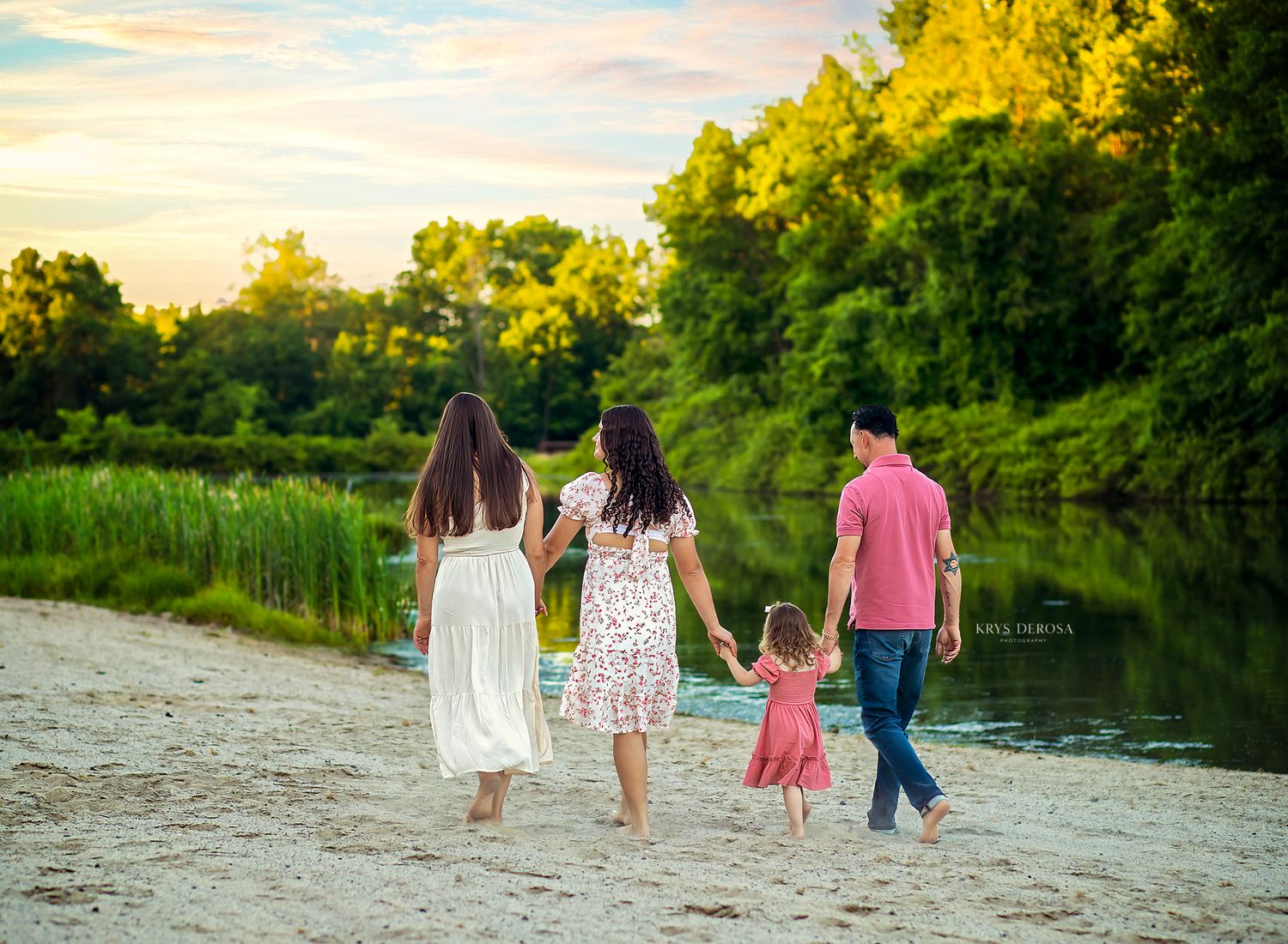 Family of four walking hand-in-hand along a sandy riverbank at sunset with lush green trees in the background.