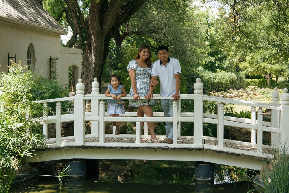 Family of three posing on a white arched garden bridge over a pond surrounded by lush green trees.