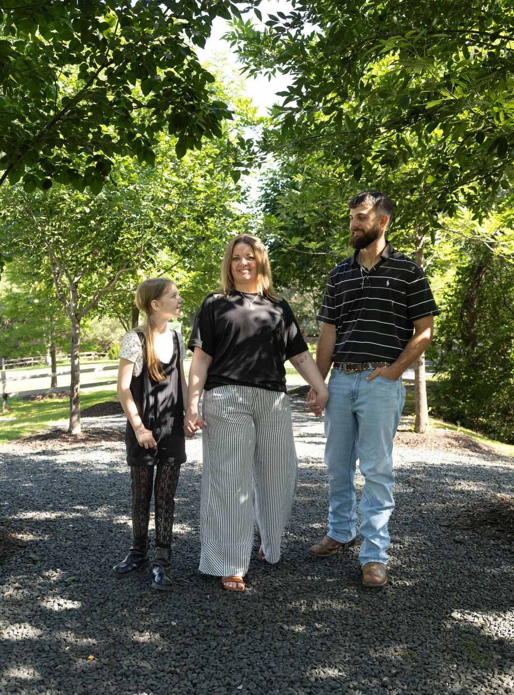 Family of three walking hand-in-hand on a gravel path surrounded by lush green trees on a sunny day.