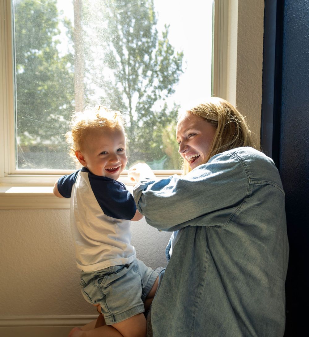 A laughing blonde woman plays with a happy red-haired toddler boy by a sunny window indoors.