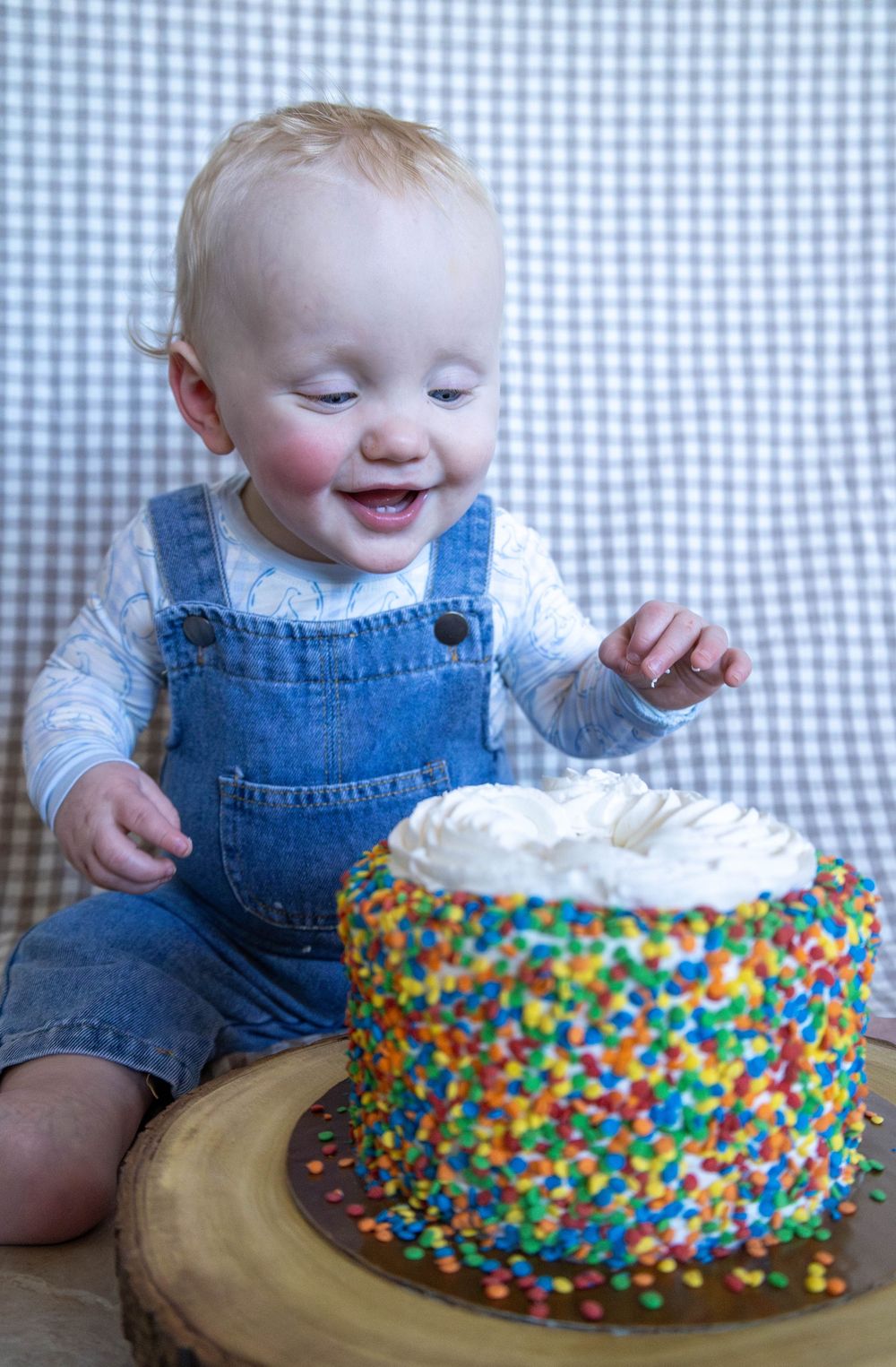 Smiling baby in denim overalls reaching toward a colorful sprinkle-covered first birthday cake on a wooden board.