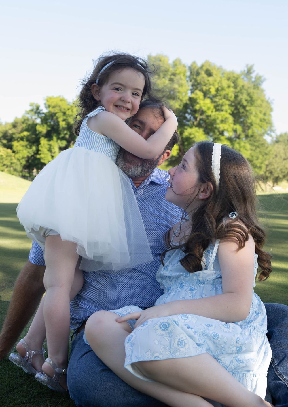 A man sits on grass hugging two young girls in white dresses outdoors near green trees.