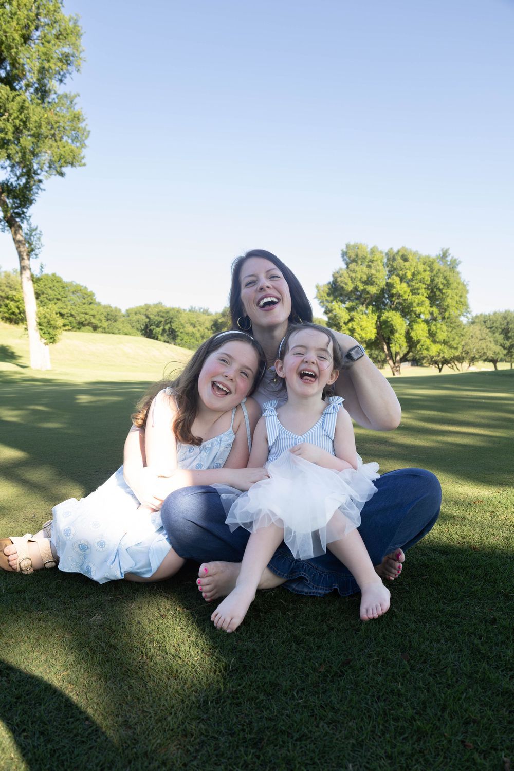 A mother and two daughters laugh joyfully while sitting together on green grass in a sunny park.