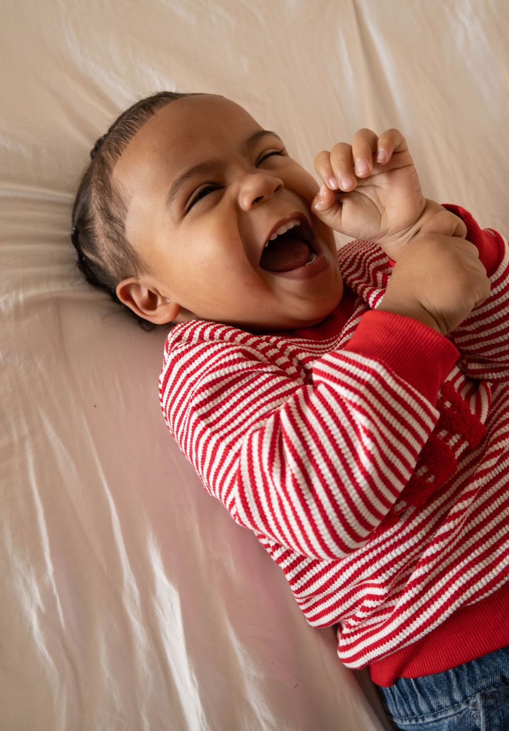 Joyful baby in red striped outfit laughing while lying on a beige surface, hands raised in delight.
