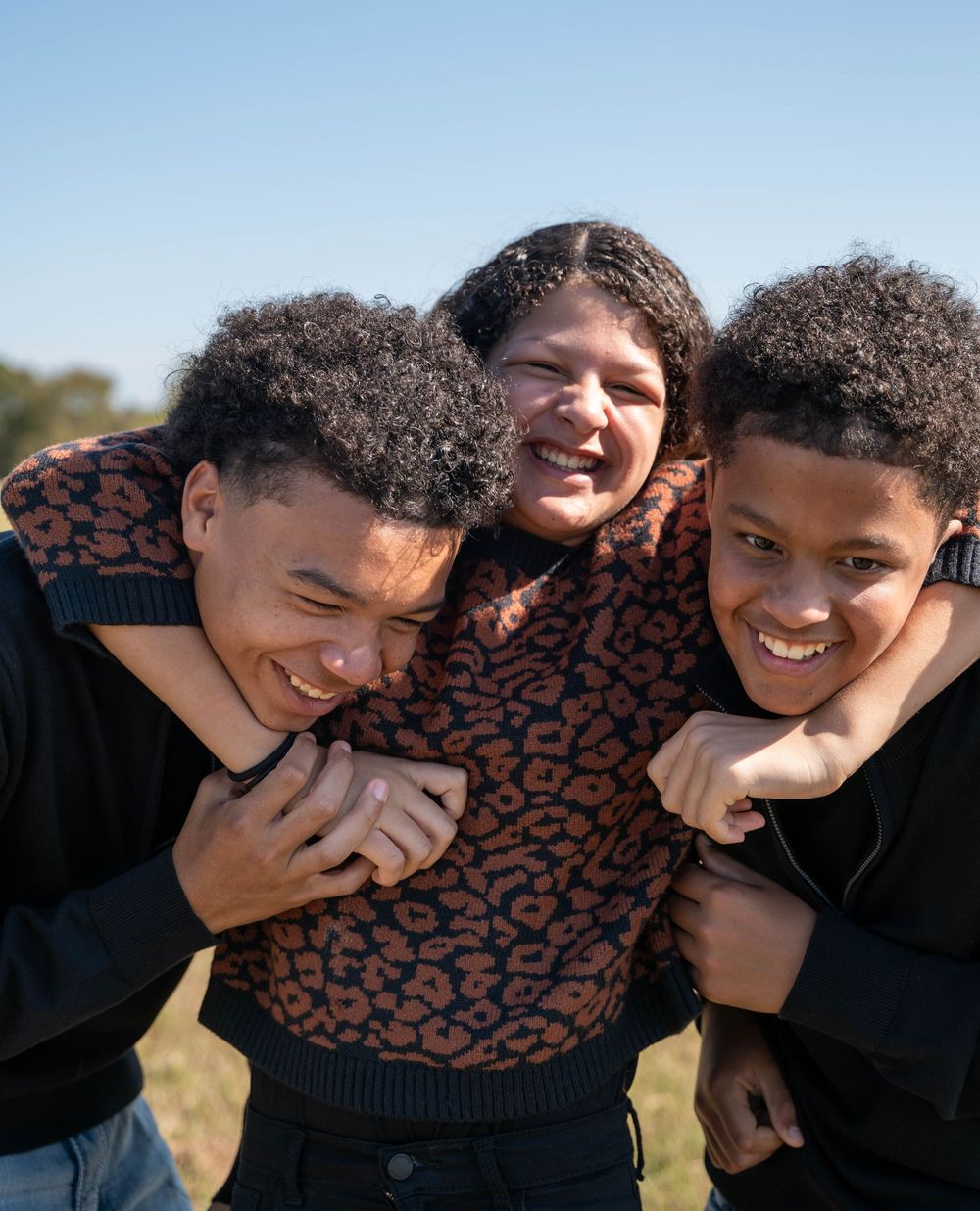 A woman smiles warmly while being hugged by two teenage boys outdoors on a sunny day.