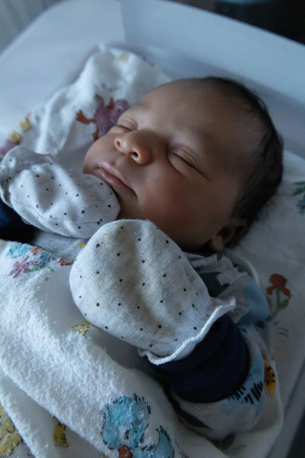 Peaceful newborn baby sleeping in a hospital bassinet, wearing white mittens and wrapped in a colorful blanket.
