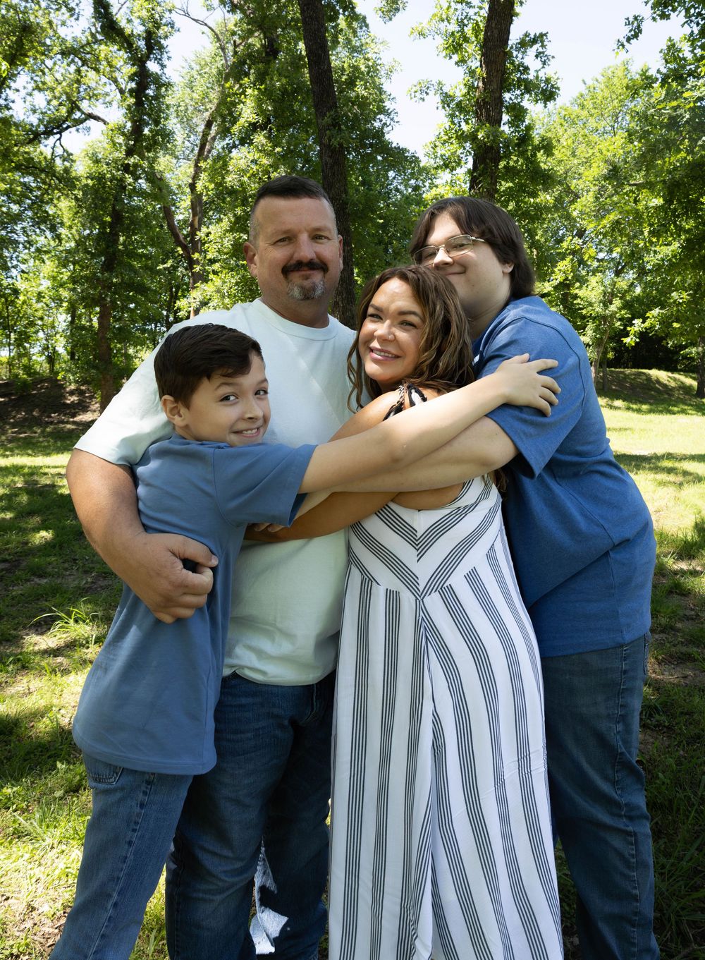 Happy family of four hugging outdoors in a lush green park setting on a sunny day.