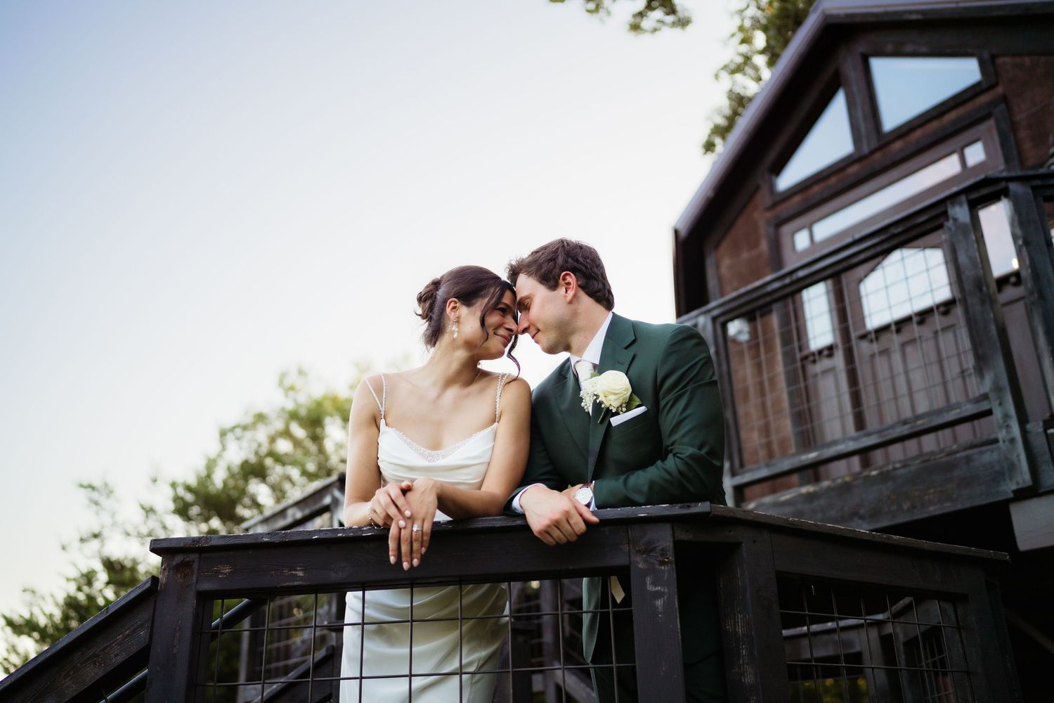 Bride and groom share a romantic kiss on a wooden balcony, with a rustic building and trees in the background.