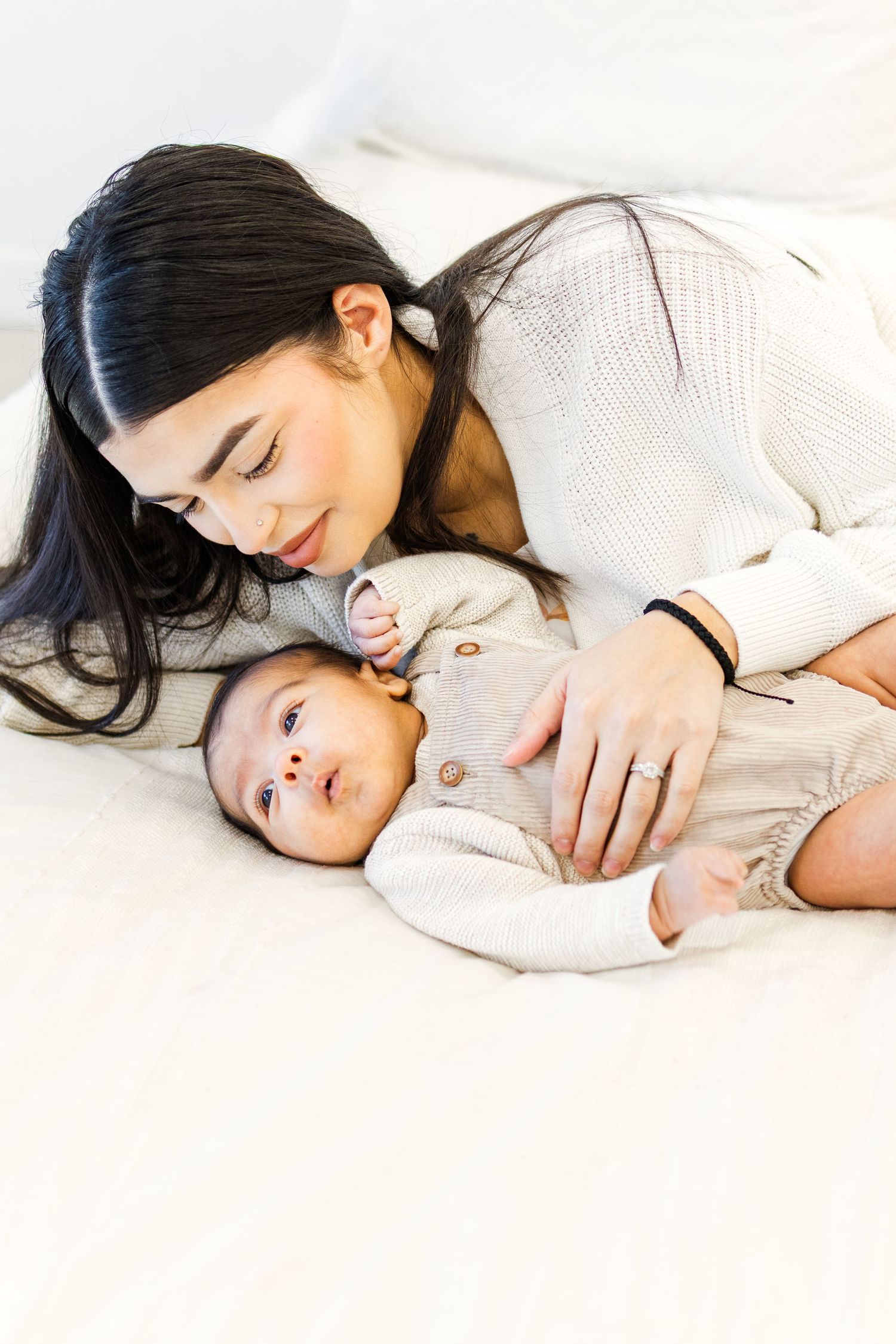 Natural light studio image in Mesa, Arizona of a young mom lying down on a bed with newborn baby, smiling down at her son
