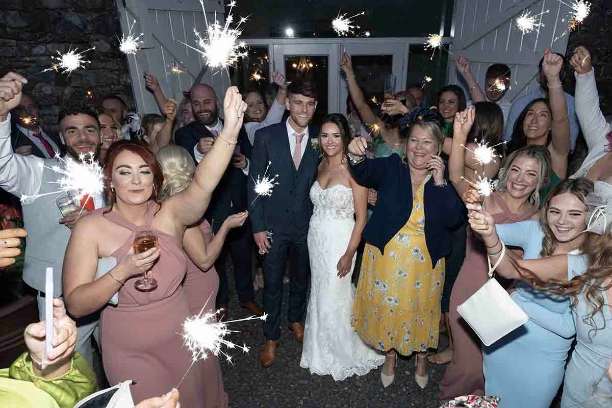 Bride and groom celebrate with wedding guests holding sparklers at a lively indoor wedding reception.