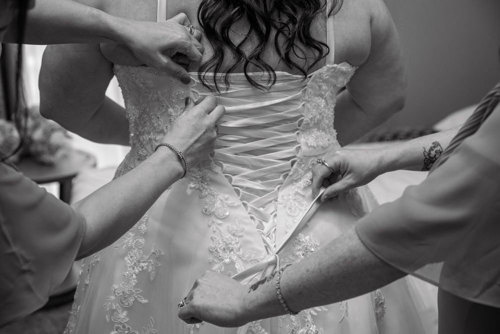 Black and white photo of bridesmaids lacing up a wedding dress corset back on a bride.
